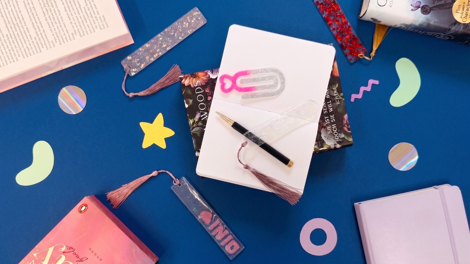 Selbstgemachte Lesezeichen aus UV-Resin mit Glitzer, Quasten und eingegossenen Motiven, arrangiert mit Büchern und einem Notizbuch