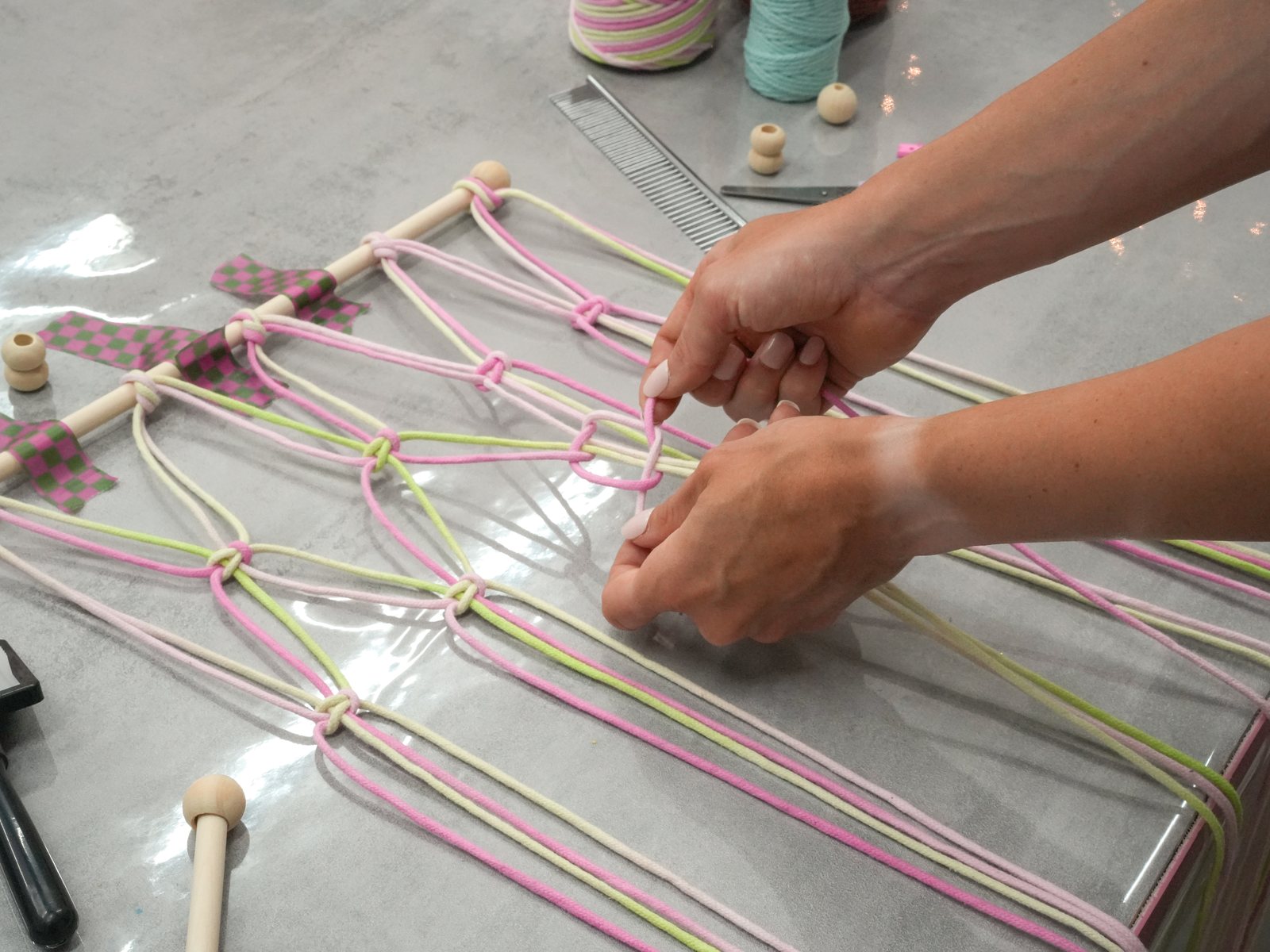 Workshop-Moment: zwei Hände knüpfen einen Knoten in pinke und limettengrüne Makramee-Schnüre, daneben Holzperlen, Kamm und Schere